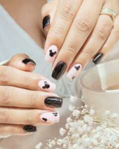 Manicure with alternating black and pale pink nails, each pink nail decorated with a black Mickey Mouse silhouette; delicate white flowers and cup in background.
