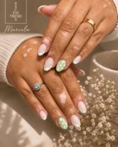 Close-up of hands showing a pastel manicure: nude nails with white French-tip accents, one green polka-dot accent nail, gold rings, and a branding watermark in the corner.