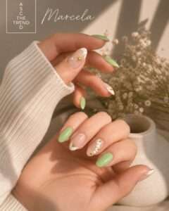 Close-up of hands with mint-green almond nails and white accent nails featuring a gold starburst and small gold beads; beige knit sleeve and soft dried flowers in the background; signature 'Marcela' visible in the upper left.