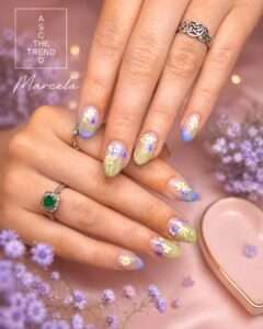 Close-up of hands with pastel floral nail art: daisies on a blue-to-green gradient, with rings on two fingers, purple background flowers visible.