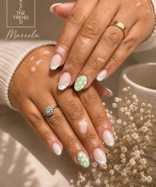 Close-up of hands showing a pastel manicure: nude nails with white French-tip accents, one green polka-dot accent nail, gold rings, and a branding watermark in the corner.